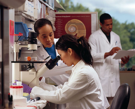 Una mujer con bata de laboratorio mirando a través de un microscopio, observada por otra mujer con bata de laboratorio. Al fondo hay un hombre.