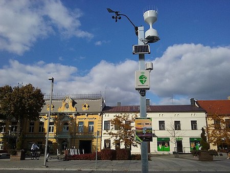 Una estación meteorológica automatizada en primer plano y casas en hilera blancas y amarillas al fondo.