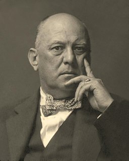 Imagen de Aleister Crowley en 1925 vistiendo un traje oscuro, camisa de vestir blanca y pajarita con la mano apoyada en la cara.