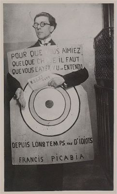 Un hombre se encuentra detrás de un cartel escrito en francés. Lleva gafas y traje, aunque apenas se le ve detrás del cartel.