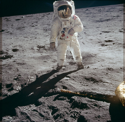 La fotografía muestra a un astronauta con un traje blanco sobre una superficie rocosa gris. El horizonte está inclinado y el fondo es negro.