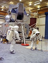 Dos astronautas practicando caminar con equipo espacial frente a una réplica de una nave espacial.