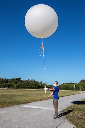 Una foto de un hombre parado en una carretera pavimentada flanqueada por campos de hierba sosteniendo la cuerda de un gran globo meteorológico blanco.