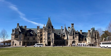 Un gran castillo gótico hecho de piedra gris y tejados altos y puntiagudos. Delante hay un césped y en la entrada hay autobuses turísticos.