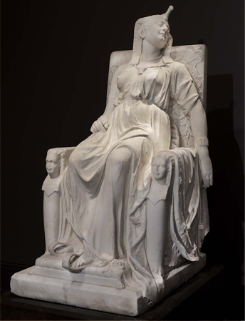 Una escultura de mármol sobre un fondo negro. La escultura representa a la reina egipcia Cleopatra aparentemente muerta en un trono.
