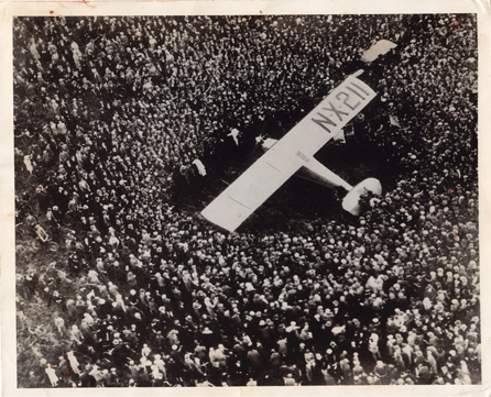 Fotografía en blanco y negro de un avión de una sola ala de los años 20 rodeado por una gran multitud. La perspectiva de la cámara es desde arriba. Matrícula de aeronave XX-211 claramente visible en el ala derecha.