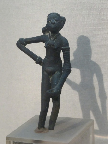 Una escultura de bronce de la civilización del valle del Indo representa a una mujer o niña joven de pie con joyas, con el brazo doblado.