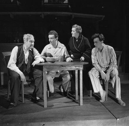 Fotografía en blanco y negro de una escena de la representación teatral de Muerte de un viajante, mostrando a cuatro actores sentados alrededor de una pequeña mesa.