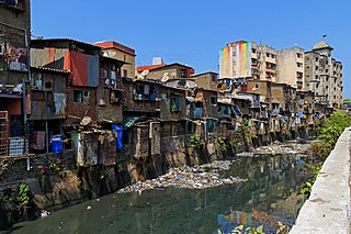 Imagen en color. La basura ensucia un canal comunitario. Junto al río, se pueden ver viviendas de tugurios con metal corrugado y rejas en las ventanas.
