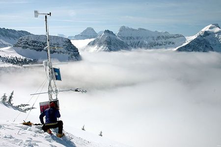 Una fotografía de una persona en la nieve junto a una estación de observación meteorológica, con picos montañosos emergiendo de una capa de nubes bajas.