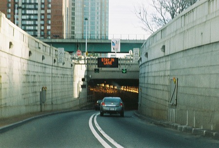 La imagen muestra un automóvil entrando por el extremo del Holland Tunnel en Nueva Jersey. Sobre la entrada del túnel hay un cartel que dice: Permanezca en el carril.