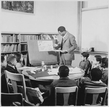 Fotografía en blanco y negro de Lawrence demostrando una lección a un grupo de niños sentados.