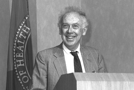 Fotografía de primer plano en blanco y negro de James Watson parado detrás de un podio.