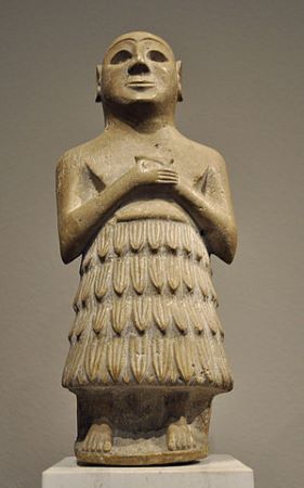 Una estatua de un hombre bien afeitado con el pecho descubierto y las manos cruzadas frente a él, vestido con una falda de plumas.