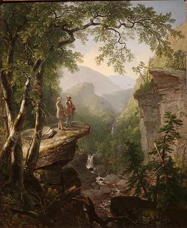 Dos hombres bien vestidos se encuentran en un acantilado con vistas a una pequeña cascada debajo con montañas al fondo.
