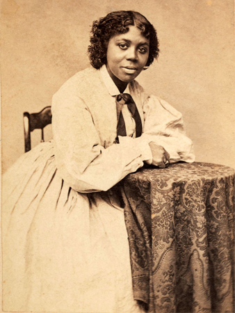 Retrato fotográfico de una mujer con atuendo del siglo XIX inclinada sobre una mesa auxiliar y mirando directamente a la cámara.