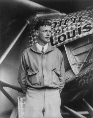 Fotografía en blanco y negro de un joven con una chaqueta de vuelo de los años 20 y corbata parado frente a un avión. Espíritu de San Luis escrito en el fuselaje.