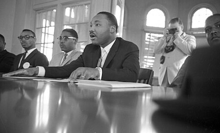 Fotografía en blanco y negro de Martin Luther King Jr. sentado en una mesa con otros cuatro defensores de los derechos civiles y fotografía de fondo.