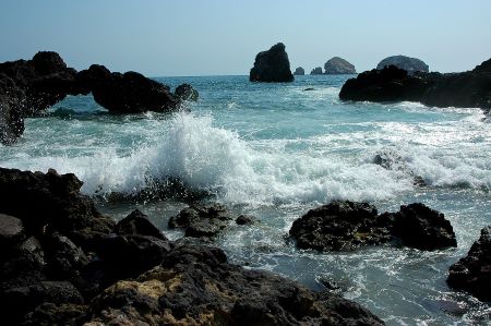 Una imagen del océano muestra olas azules y blancas rompiendo contra rocas oscuras.