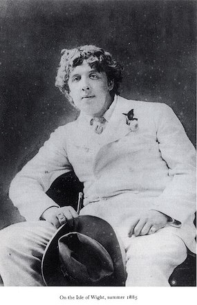 Fotografía en blanco y negro de Oscar Wilde, sentado, vestido con traje blanco y sombrero en mano.