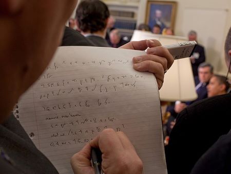 Vista por encima del hombro de las notas del periodista en taquigrafía. Obama al fondo, borroso.