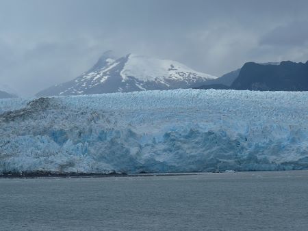 Una fotografía en color con el mar en primer plano, una masa puntiaguda de hielo en el centro y montañas nevadas al fondo.
