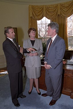 Imagen en color de Janet Reno con un traje de falda gris y camisa negra en la oficina oval con Joe Biden y Bill Clinton.