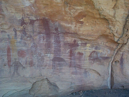 Una pared de roca de color naranja y tostado presenta pinturas de figuras humanas simplificadas y formas abstractas en pintura roja.