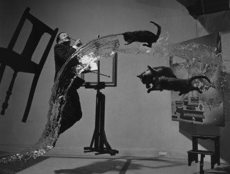 Foto en blanco y negro. El hombre salta a la izquierda, junto a una silla en el aire. Tres gatos están en el aire a la derecha.