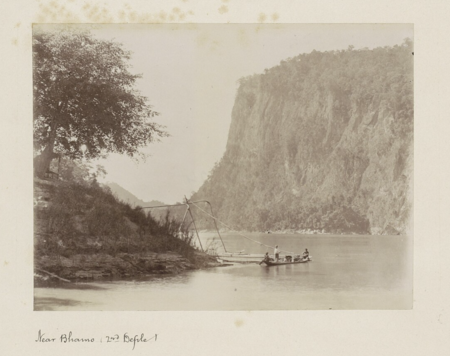 Una foto antigua en tonos sepia que representa un pequeño barco a lo largo de la orilla del río Irrawaddy, con el segundo desfiladero al fondo.