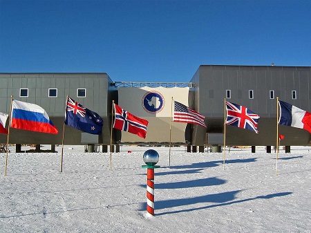 La imagen muestra el edificio de la estación de investigación, varias banderas y el Polo Sur ceremonial.