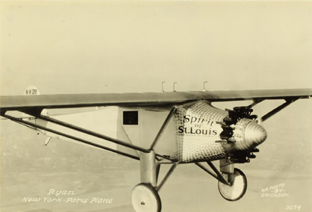 Foto en tonos sepia de un avión de una sola ala volando. No se ve ningún parabrisas. Se ve una única ventana lateral. En el fuselaje está escrito Spirit of St. Louis.