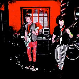 Una foto con un fondo rojo vivo contrastado con colores muy oscuros. Dos músicos actúan en el escenario.