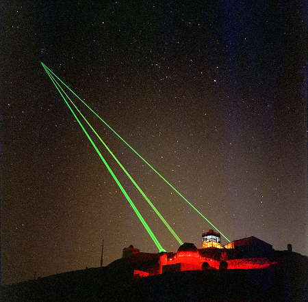 La imagen muestra tres láseres verdes de una instalación dirigidos hacia arriba y enfocados en un punto del cielo.