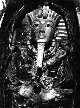 Una fotografía en blanco y negro de la máscara de Tutankamón in situ, se ve suciedad a su alrededor. La cobra erguida adorna su tocado.