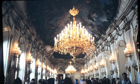Una gran sala ornamentada con 2 grandes lámparas de araña. Las paredes son blancas con dorados dorados, espejos a un lado y ventanas al otro.