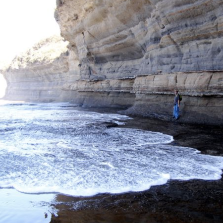 Una persona se encuentra junto a un acantilado marrón junto a la playa con capas de roca sedimentaria visibles mientras la espuma blanca del mar cubre el suelo